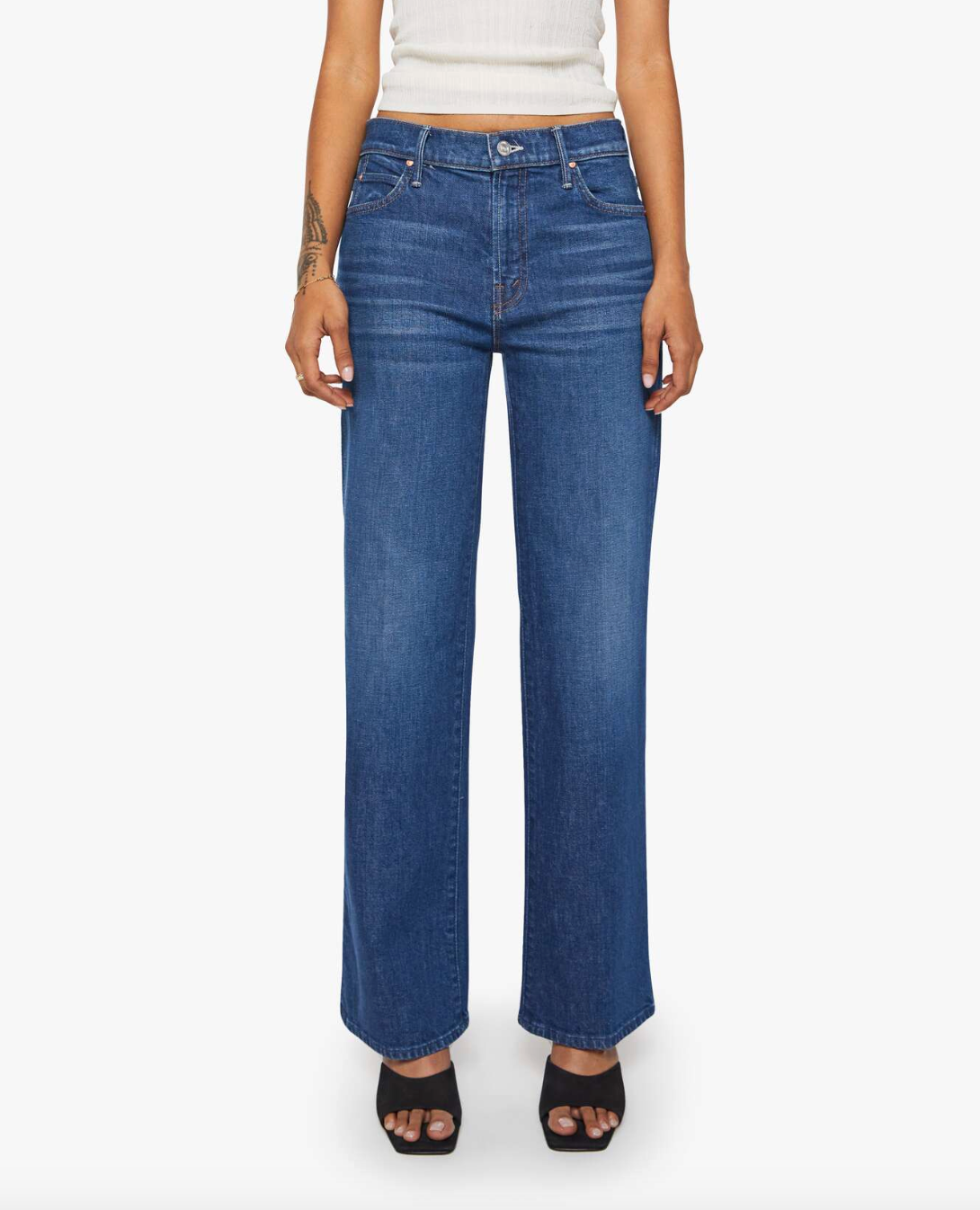 A person wears Mother’s Lil Kick It high-waisted, wide-leg blue jeans—crafted for petites with front and back pockets—paired with a white sleeveless top and black open-toe heels, standing against a white background.