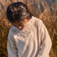 A young girl with dark hair looks down while standing outdoors in tall, dry grass. She wears the Girls’ Osalia Sweatshirt by Louise Misha, a cream-colored fleece featuring delicate embroidery and lace detail around the collar.
