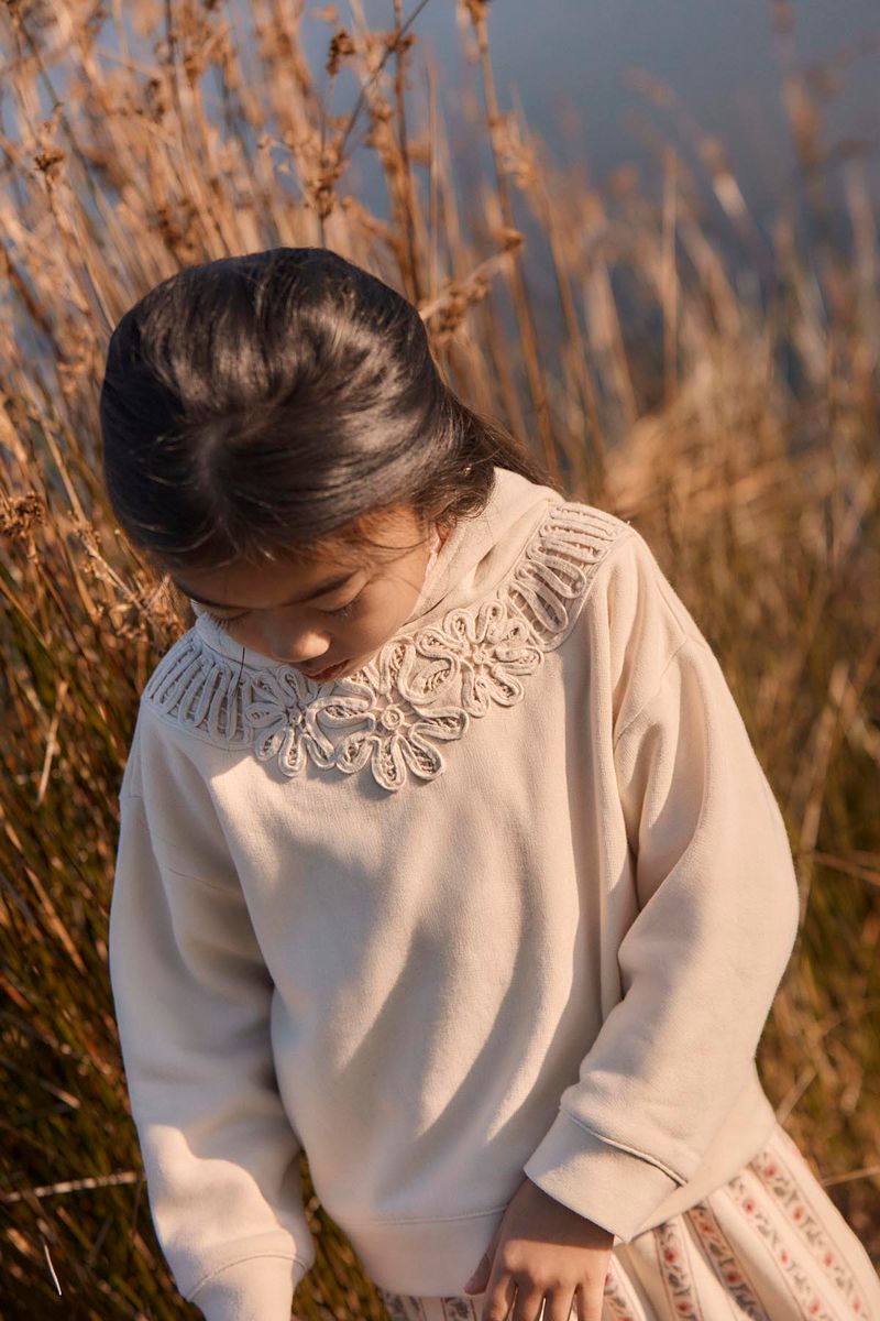 A young girl with dark hair looks down while standing outdoors in tall, dry grass. She wears the Girls’ Osalia Sweatshirt by Louise Misha, a cream-colored fleece featuring delicate embroidery and lace detail around the collar.