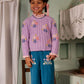 A young girl stands on a wooden chair indoors, smiling with ribbons in her hair, wearing the Louise Misha Girls’ Zuzanna Sweater—a lavender knit style featuring handmade floral embroidery and a crocheted scalloped neckline—paired with teal pants embellished with flower embroidery.