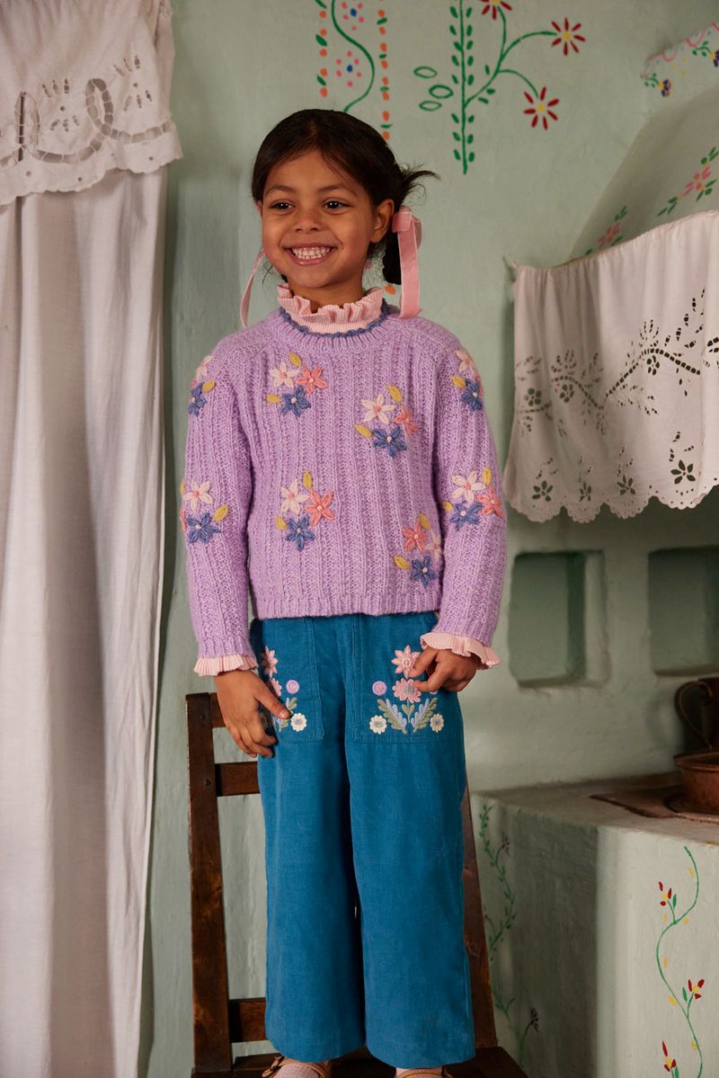 A young girl stands on a wooden chair indoors, smiling with ribbons in her hair, wearing the Louise Misha Girls’ Zuzanna Sweater—a lavender knit style featuring handmade floral embroidery and a crocheted scalloped neckline—paired with teal pants embellished with flower embroidery.