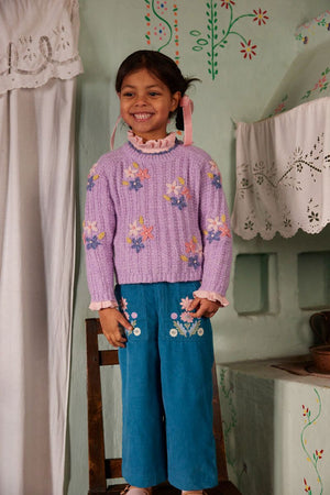 A young girl stands on a wooden chair indoors, smiling with ribbons in her hair, wearing the Louise Misha Girls’ Zuzanna Sweater—a lavender knit style featuring handmade floral embroidery and a crocheted scalloped neckline—paired with teal pants embellished with flower embroidery.