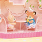 Two Calico Critters animal figurines, a mouse and a deer, sit together at a table inside the Baby Dream Amusement Park playset, featuring a charming pink, dollhouse-style setting.