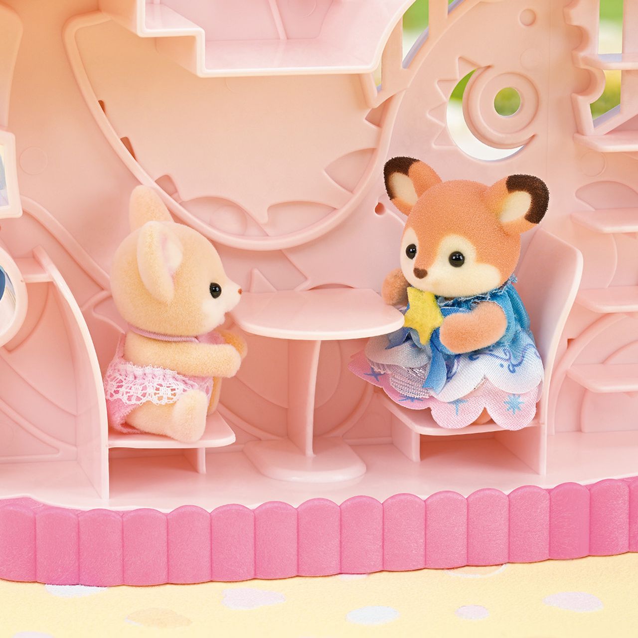 Two Calico Critters animal figurines, a mouse and a deer, sit together at a table inside the Baby Dream Amusement Park playset, featuring a charming pink, dollhouse-style setting.