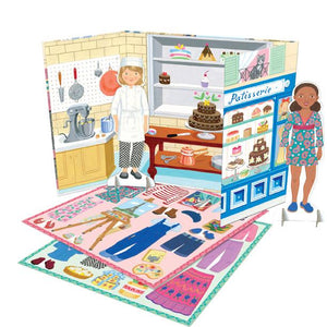 Two Eeboo Paper Doll Set figures stand in a bakery-themed playset, complete with shelves of cakes, counters, and a floor scattered with outfits and accessories for dressing the dolls—making it ideal for imaginative play at home or as a portable travel toy.