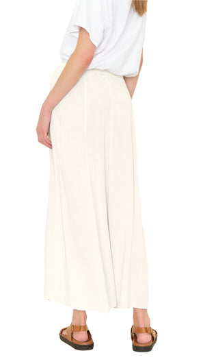A person relaxedly stands in sandals, wearing a white T-shirt and Xirena's Deon Skirt, featuring an elasticized waist. One hand rests by their side while the skirt’s flowy cotton gauze adds elegance.