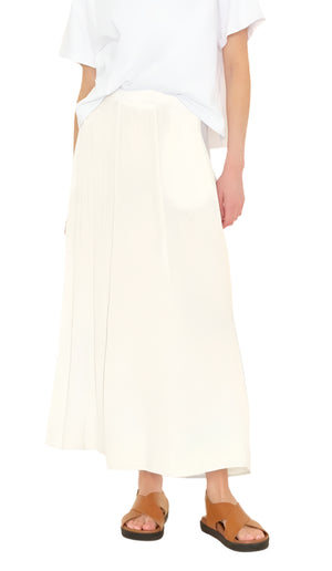 Against a white background, someone wears a white t-shirt, the Deon Skirt made from cotton gauze by Xirena, and brown sandals. The skirt's elasticized waist enhances both comfort and style.