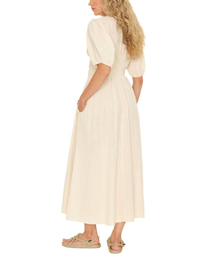 A woman stands slightly turned away from the camera in the Xirena Lawsyn Dress, a light beige, short-sleeve cotton gauze midi, paired with sandals against a plain background.