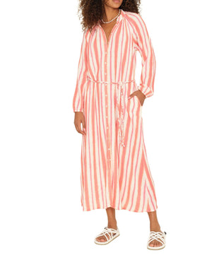 A woman wears the Alicia Dress by Xirena, a long-sleeve cotton gauze midi with pink and white vertical stripes, button front, waist tie, and pairs it with white sandals.