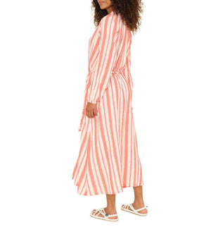 A woman stands sideways against a white background wearing the Xirena Alicia Dress, a long-sleeved pink and white striped cotton gauze midi dress with a tie waist, paired with white sandals.