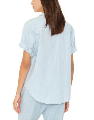 A person is seen from the back wearing the Xirena Channing Chambray Shirt in light blue cotton, styled with rolled cuffs and matching pants—a versatile essential for any wardrobe.