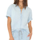 A person wears the Xirena Channing Chambray Shirt in lightweight cotton with rolled sleeves, paired with matching light blue drawstring pants, standing against a plain background.