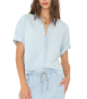 A person wears the Xirena Channing Chambray Shirt in lightweight cotton with rolled sleeves, paired with matching light blue drawstring pants, standing against a plain background.