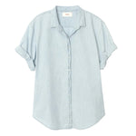 The Channing Chambray Shirt by Xirena is an essential, lightweight cotton top in light blue, featuring short rolled sleeves, a collar, and a curved hem, shown against a plain white background.