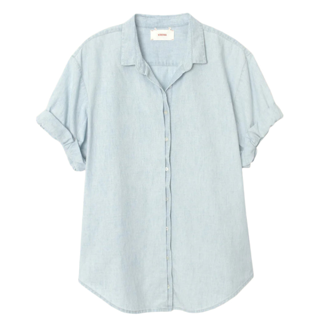 The Channing Chambray Shirt by Xirena is an essential, lightweight cotton top in light blue, featuring short rolled sleeves, a collar, and a curved hem, shown against a plain white background.