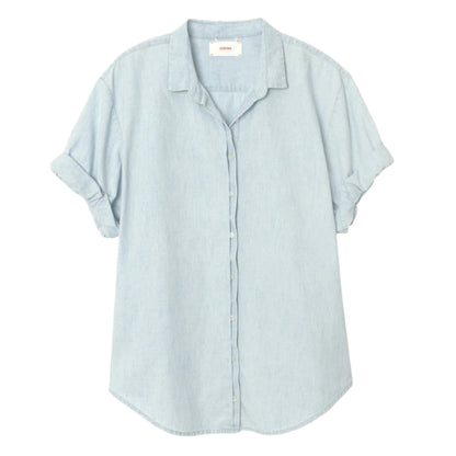 The Channing Chambray Shirt by Xirena is an essential, lightweight cotton top in light blue, featuring short rolled sleeves, a collar, and a curved hem, shown against a plain white background.
