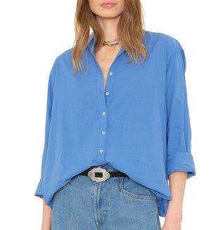 A woman wears the Xirena Beau Shirt by Xirena, a loose blue cotton poplin menswear-inspired piece with rolled sleeves, styled with blue jeans, a decorative belt, and a multicolored necklace. Her hair is down and face partially out of frame.