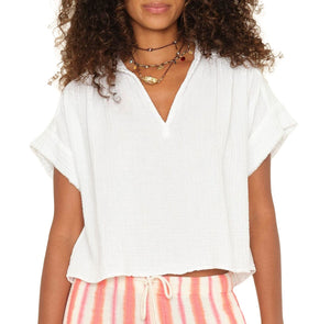 A woman wears the Xirena Reggie Top, a white cotton gauze pullover with short sleeves and a V-neck, paired with striped drawstring shorts, shown from shoulders to hips.