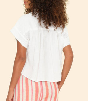 A woman with curly hair is shown from the back, wearing the Xirena Reggie Top in white cotton gauze and pink-and-white striped shorts against a beige background.