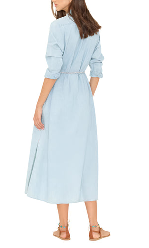 A woman faces away, wearing the Xirena Bowen Dress in a light blue wash chambray with rolled-up sleeves, a belted waist, and tan flat sandals.