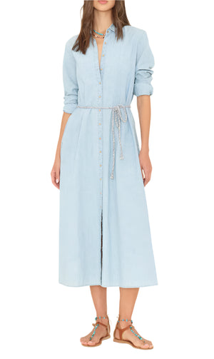 A woman wears the Xirena Bowen Dress, a light blue chambray button-down midi with long sleeves and a tie belt, paired with brown sandals. Her head is partially out of frame.