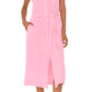 A person wears the Xirena Aliza Dress—a light pink, button-front midi in double-faced cotton gauze with adjustable tie-belt and side pockets—paired with strappy flat sandals.