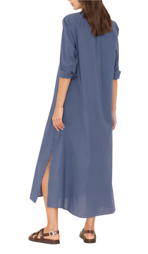 A woman is shown from the back wearing the Xirena Boden Dress, a loose-fitting blue shirtdress with rolled-up sleeves and side slits, paired with brown sandals.