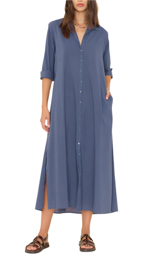 A person wearing the Xirena Boden Dress—a blue, long-sleeve, button-front cotton poplin midi shirtdress with side slits—styled with brown sandals and layered necklaces, standing with hands in pockets.