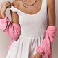 A woman wears the Xirena Florence Dress, a white sleeveless midi sundress with a smocked bodice, layered pink necklaces, and a pink shirt draped over her shoulders.
