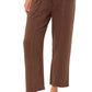 A person wears the Xirena Ozzie Pant—loose, brown, ankle-length cotton gauze pants with an elastic drawstring waist—paired with a white top and tan strappy sandals.