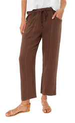 A person wears the Xirena Ozzie Pant—loose, brown, ankle-length cotton gauze pants with an elastic drawstring waist—paired with a white top and tan strappy sandals.