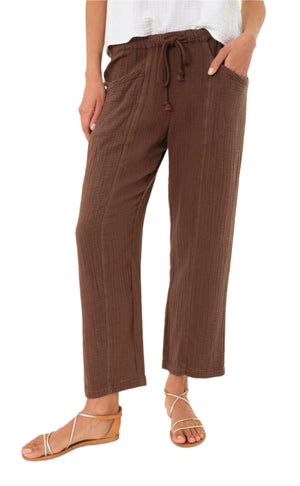 A person wears the Xirena Ozzie Pant—loose, brown, ankle-length cotton gauze pants with an elastic drawstring waist—paired with a white top and tan strappy sandals.