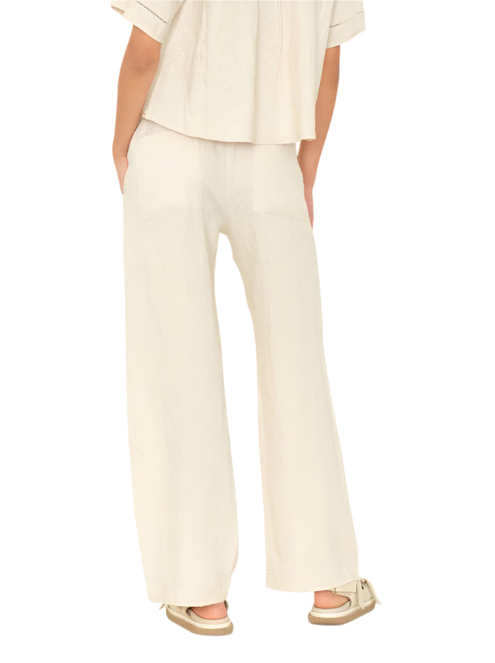 Model shown from the back wearing Xirena's Romany Pant in a loose, cream hue with an elastic waist, paired with a matching short-sleeve top and beige sandals, set against a plain background.