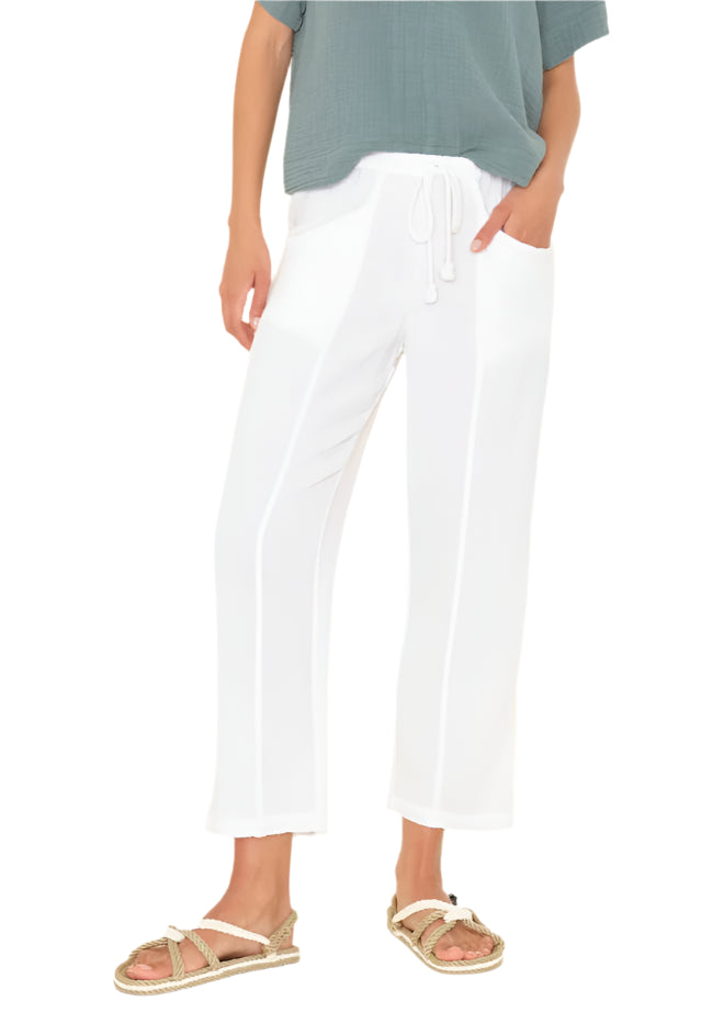 A person wearing a green short-sleeve top, Xirena Ozzie Pant—cotton gauze cropped pull-on style with an elasticized drawstring waist—and beige sandals stands with one hand in their pocket.
