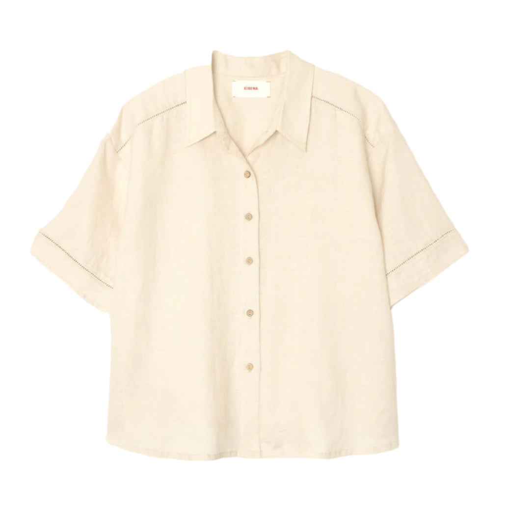 The Xirena Hendry Shirt is a beige, short-sleeved linen button-up with coconut buttons and a collar, shown on a plain white background.