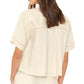 A woman is seen from the back wearing the Xirena Hendry Shirt—an off-white, short-sleeve, loose-fit linen top with subtle stitching and coconut buttons.