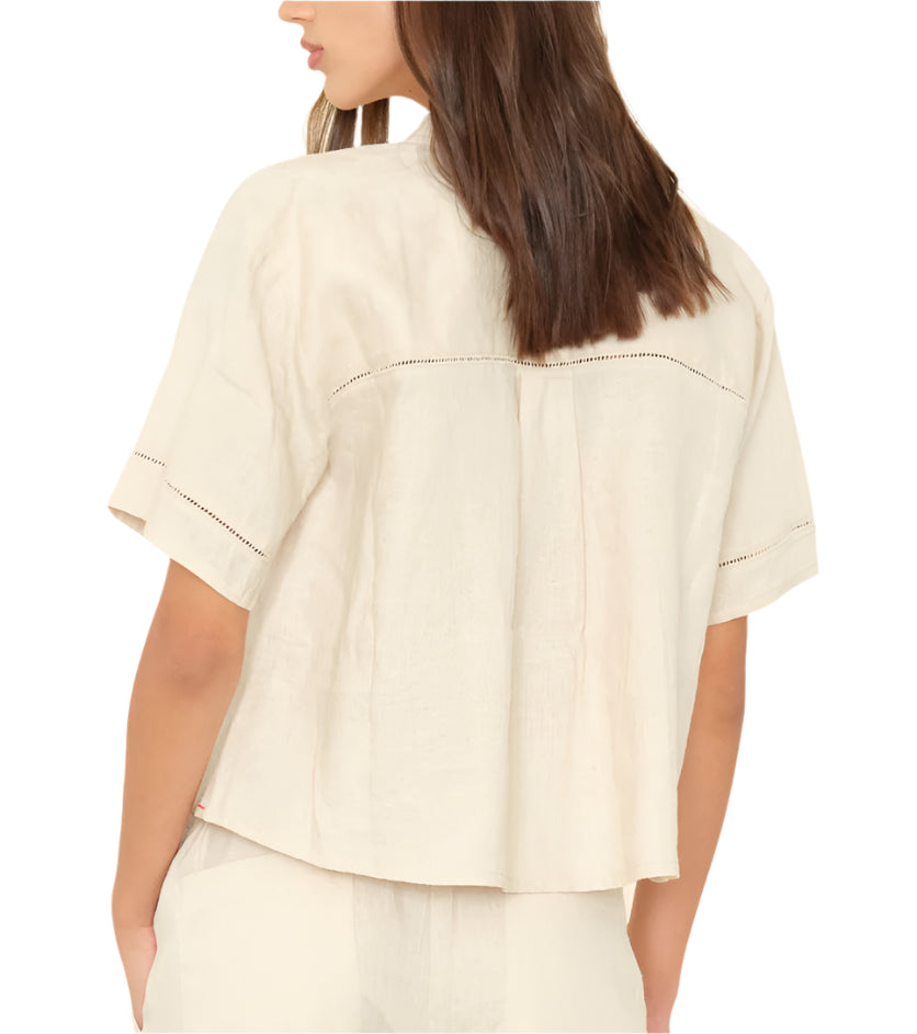 A woman is seen from the back wearing the Xirena Hendry Shirt—an off-white, short-sleeve, loose-fit linen top with subtle stitching and coconut buttons.