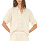 A woman in the Xirena Hendry Shirt, a light beige linen button-front top with coconut buttons and collar, paired with matching elastic-waist pants, stands hands on hips.