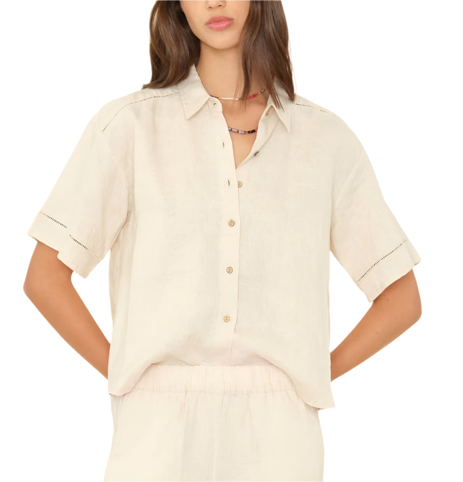 A woman in the Xirena Hendry Shirt, a light beige linen button-front top with coconut buttons and collar, paired with matching elastic-waist pants, stands hands on hips.