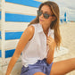 A woman wearing sunglasses, a white sleeveless shirt, and Xirena’s Declan Short—blue with an elasticized drawstring waist—sits on the sand beside a white and blue striped beach hut.