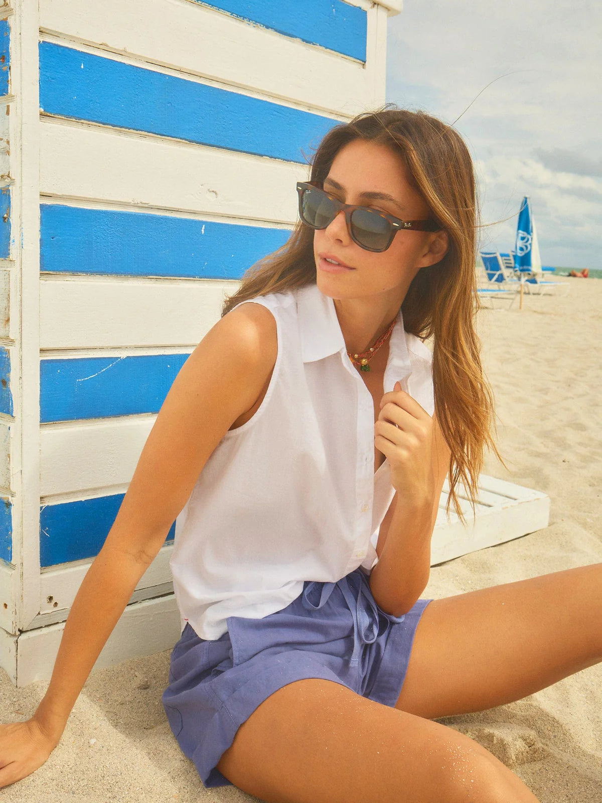 A woman wearing sunglasses, a white sleeveless shirt, and Xirena’s Declan Short—blue with an elasticized drawstring waist—sits on the sand beside a white and blue striped beach hut.