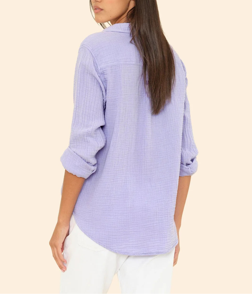 A woman with long brown hair, wearing the Xirena Scout Shirt in light purple with rolled-up sleeves and white pants, stands facing away and looks over her shoulder.