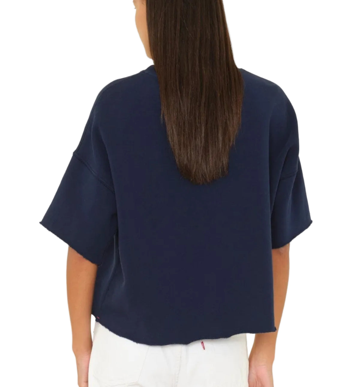 A person facing away from the camera against a white background is wearing the Xirena Romeo Sweatshirt Tee—a loose-fitting, boxy navy blue top with raw-edge sleeves—paired with white pants.