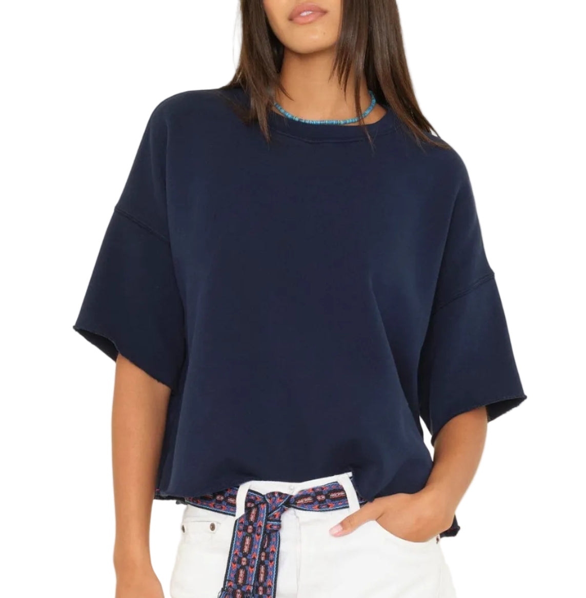 A woman wearing the Xirena Romeo Sweatshirt Tee—a loose, navy blue short-sleeve top with a boxy fit—styled with white pants, a blue beaded necklace, and a patterned belt, stands with her left hand in her pocket.