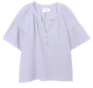 The Nia Top by Xirena is a light purple cotton gauze popover with short flutter sleeves, a round collar, button placket, and loose-fitting textured fabric.
