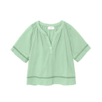 The Sadie Top by Xirena is a light green women’s blouse made from soft cotton poplin, featuring a relaxed fit, round neckline, and partial henley button-front.