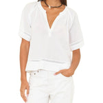 A woman wears the Xirena Sadie Top, a loose-fitting white cotton poplin short-sleeve blouse with a V-neck, styled with white pants and a black choker necklace.