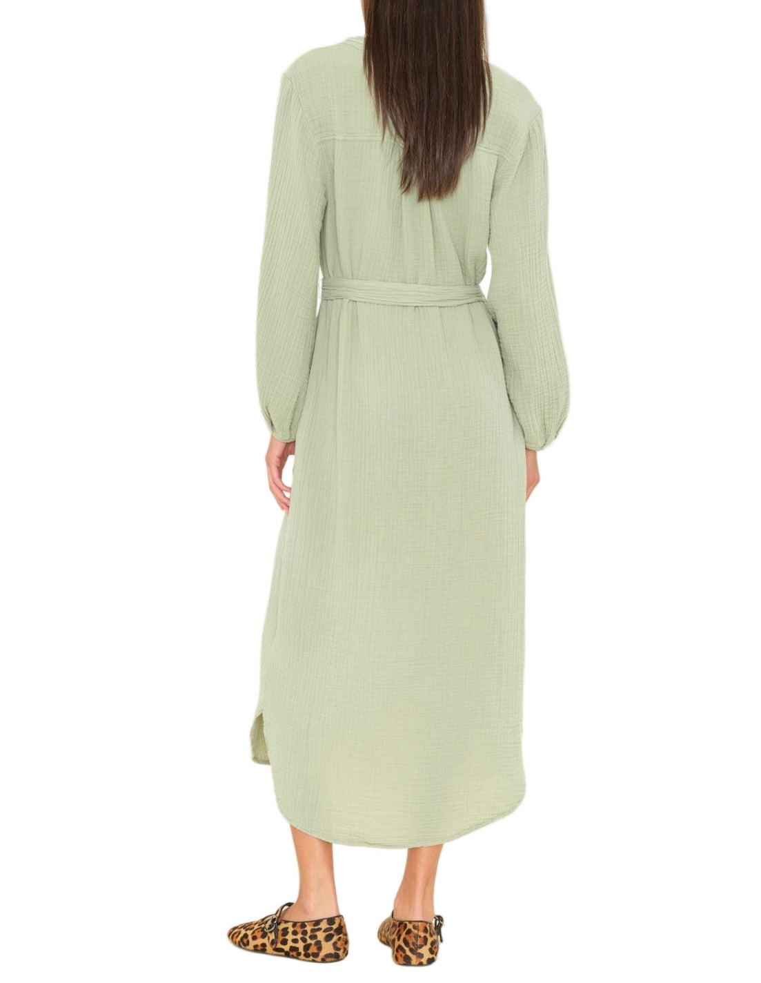 A woman stands facing away, wearing the Xirena Josephine Dress in light green washed cotton gauze with blouson sleeves and a belt, paired with leopard print loafers.