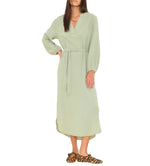 A woman with long brown hair faces forward, wearing the Xirena Josephine Dress in light green washed cotton gauze with blouson sleeves and leopard print slippers.