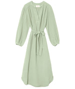 The Josephine Dress by Xirena is a light green V-neck midi with blouson sleeves, waist tie, and relaxed fit in soft cotton gauze. Knee-length style shown on a white background.